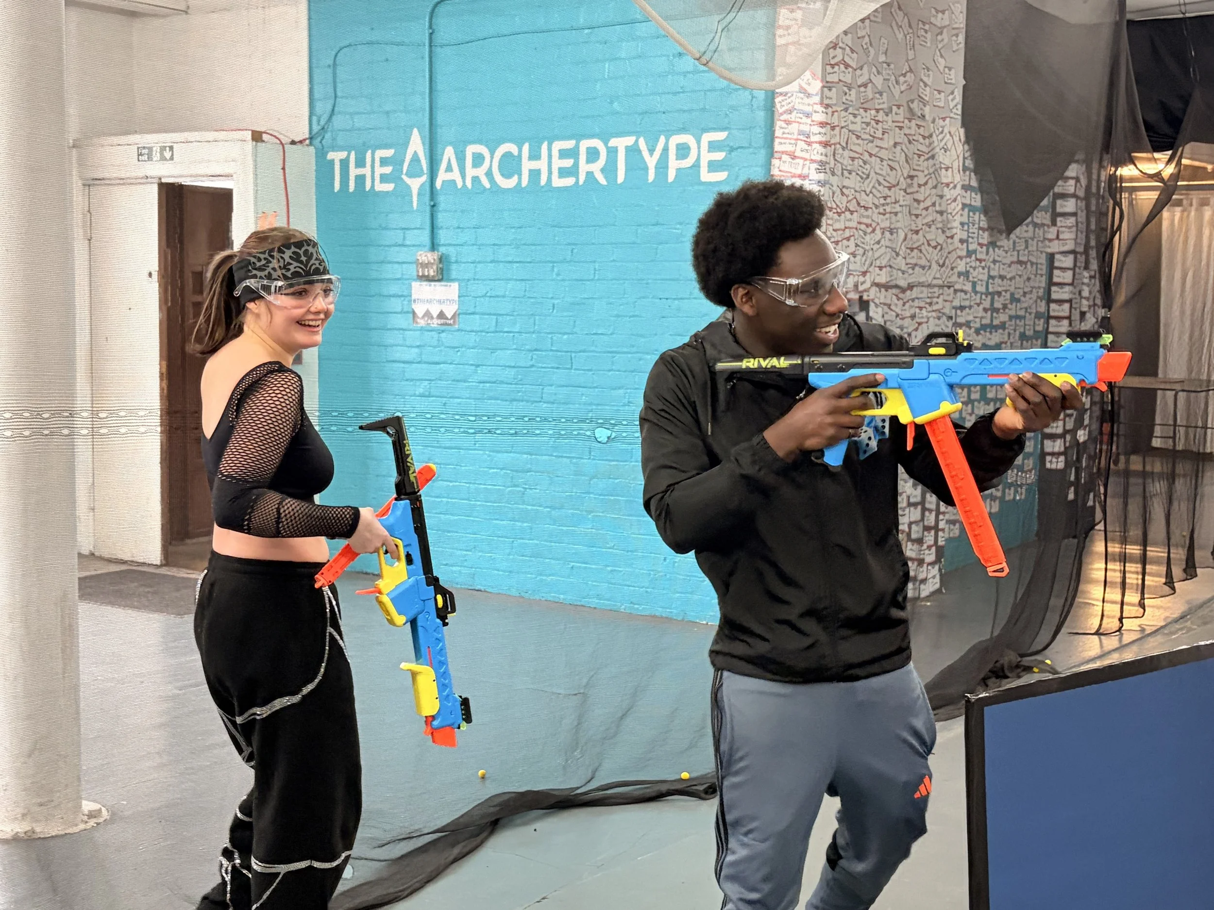 Two people wearing safety glasses aim foam dart guns in an indoor space with the sign "The Archetype" on the blue wall behind them.