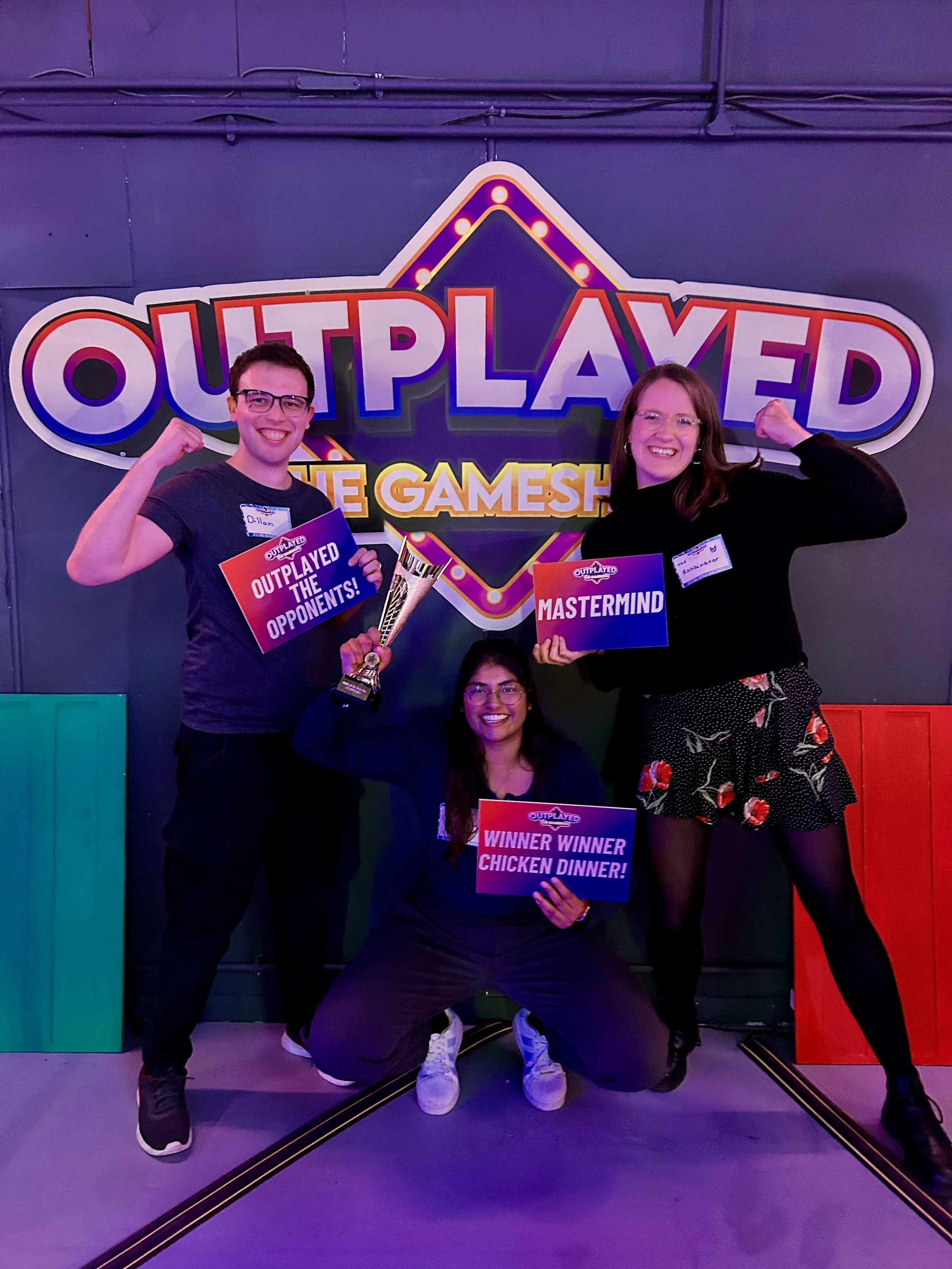 Three people celebrating at an event called OUTPLAYED THE GAMESHOW, holding signs that read "OUTPLAYED THE OPPONENTS!", "MASTERMIND," and "WINNER WINNER CHICKEN DINNER!" in front of a colorful backdrop.