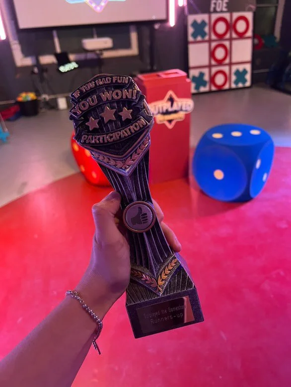 Hand holding a trophy with a bow tie and thumbs-up sticker, in a game room with giant dice and a tic-tac-toe board in the background.