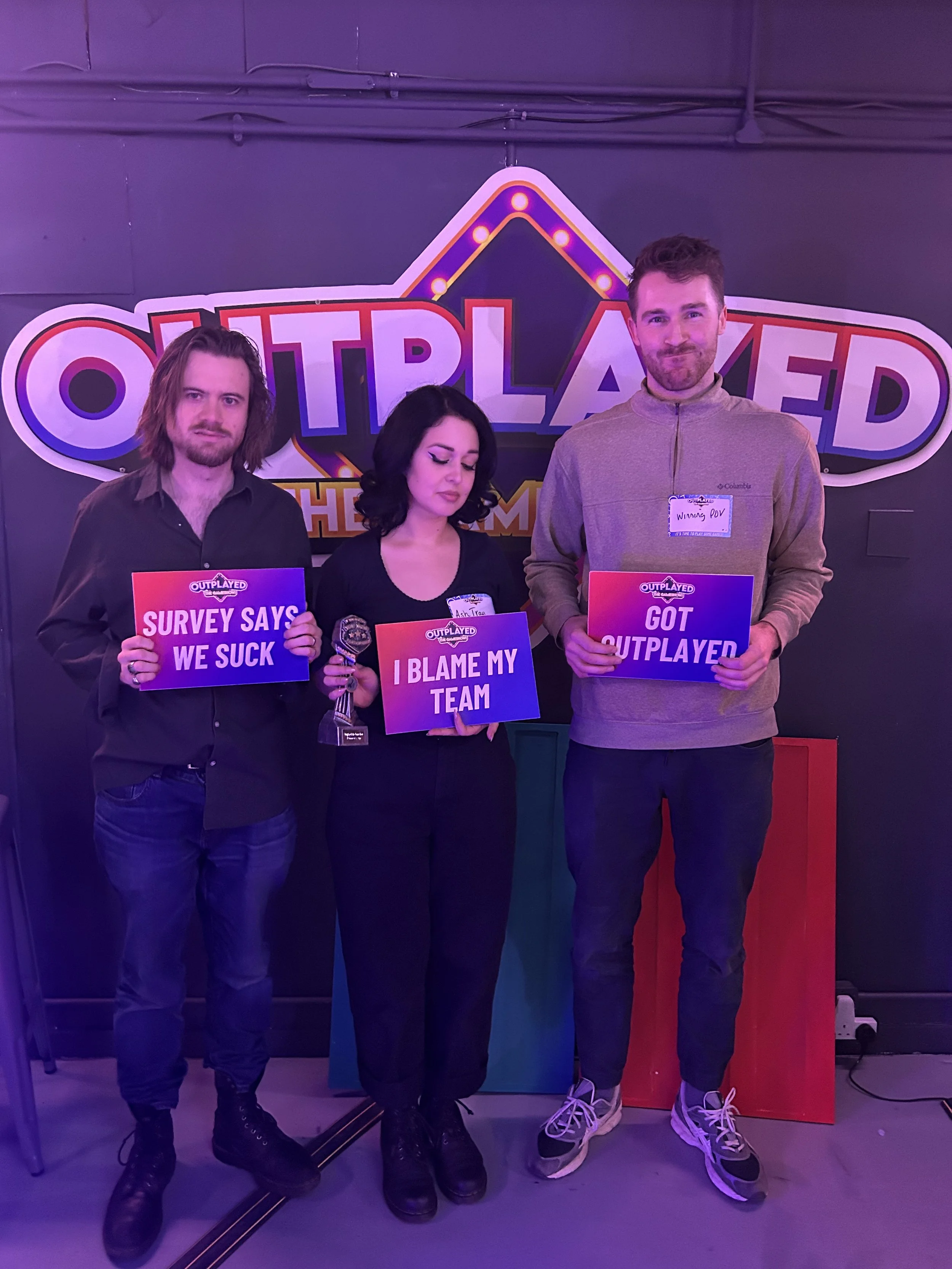 Three people standing in front of a sign that reads "Outplayed"; they are holding signs with humorous messages related to a game show or quiz competition.