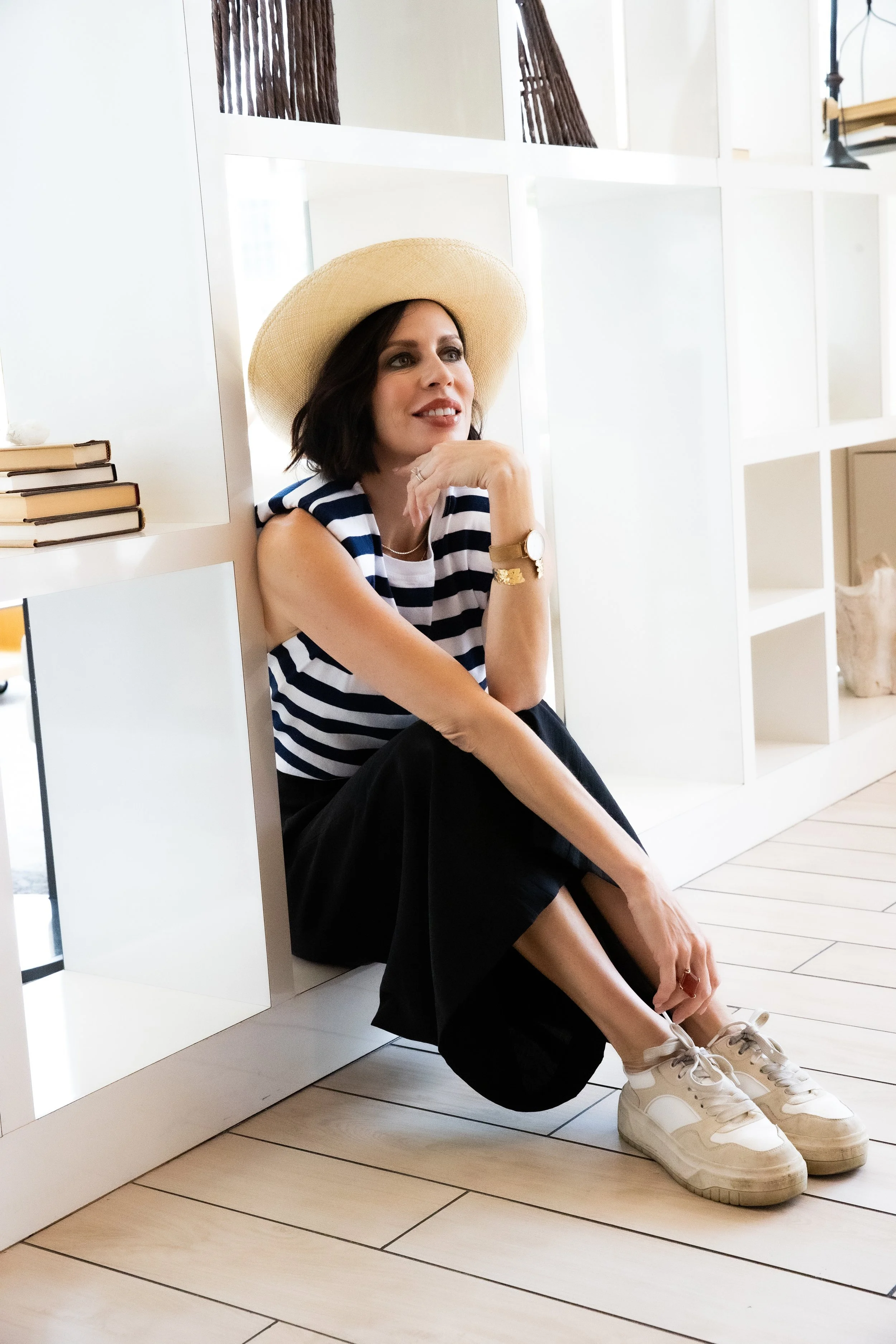 A woman with dark hair wearing a wide-brimmed hat and striped top sitting on the floor next to white shelving units, smiling and looking to her left.