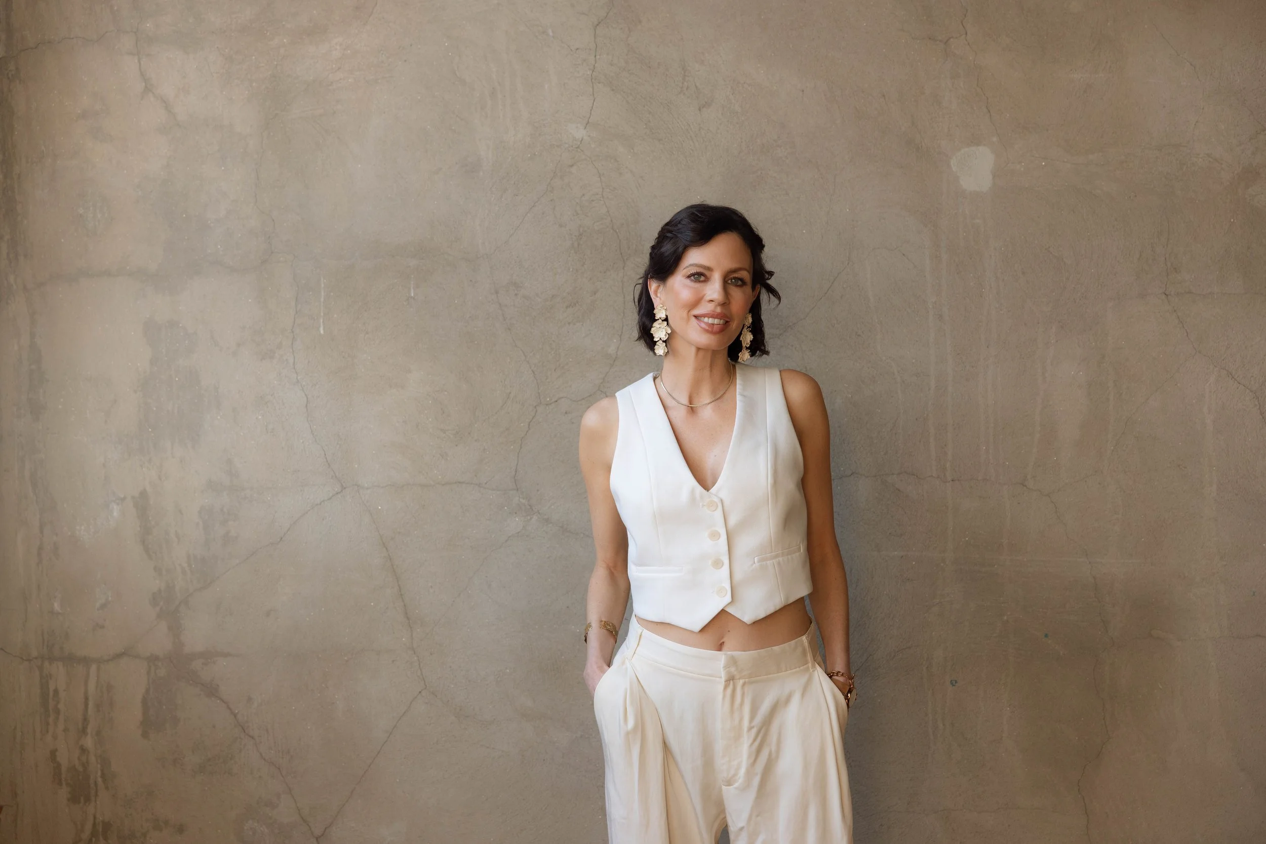 A woman with dark hair wearing a white sleeveless vest and cream-colored pants standing against a textured beige wall.