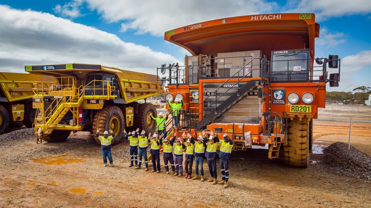 Group of construction workers gathered in front of large orange and yellow mining dump trucks at a construction site, with some workers standing on the trucks' steps, all wearing safety helmets and reflective vests.