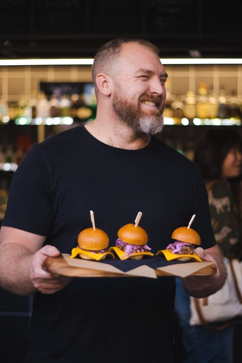A man with a beard wearing a black t-shirt smiling and holding a wooden tray with three mini cheeseburgers with purple coleslaw and yellow cheese, set in a restaurant or bar.