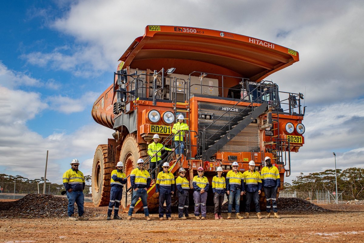 Group of workers in yellow and blue safety uniforms and white helmets standing in front of a large orange mining dump truck at a construction site.