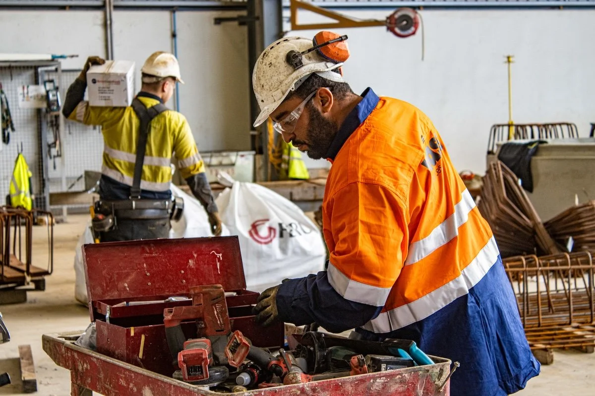 Construction workers in safety gear working in a workshop, with one worker examining tools in a red toolbox.