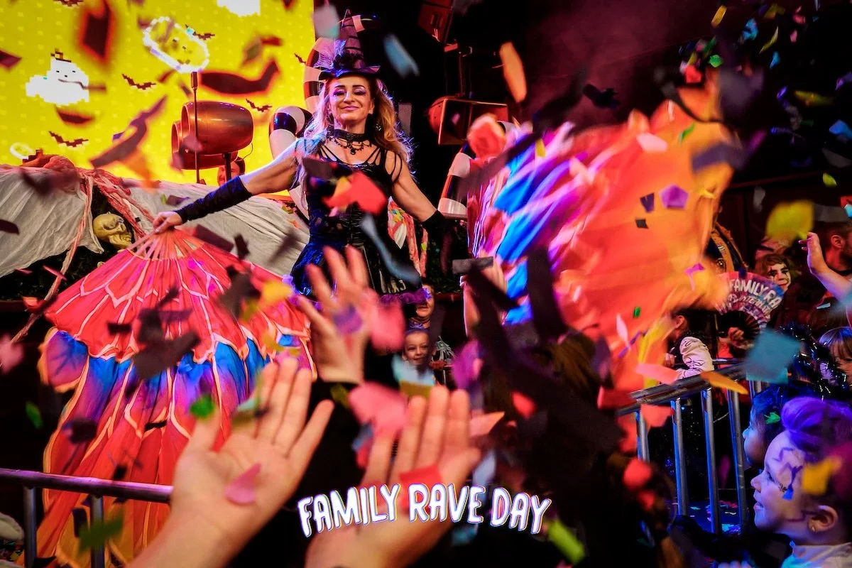 People celebrating at a Halloween-themed event called Family Rave Day, with a woman dressed as a witch, colorful costumes, confetti in the air, and a decorated background with jack-o'-lantern faces.
