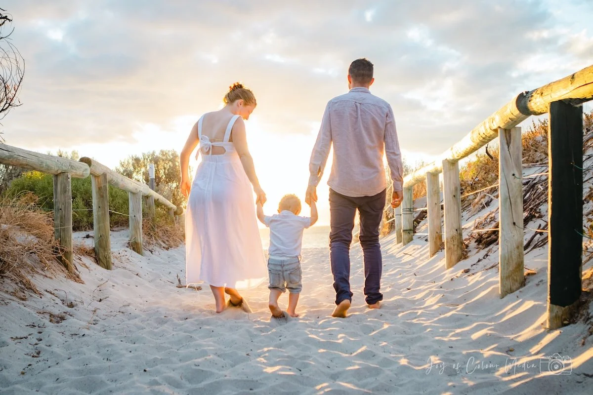 A family of four walking hand in hand on a sandy beach at sunset, with a wooden fence on either side and trees in the background.