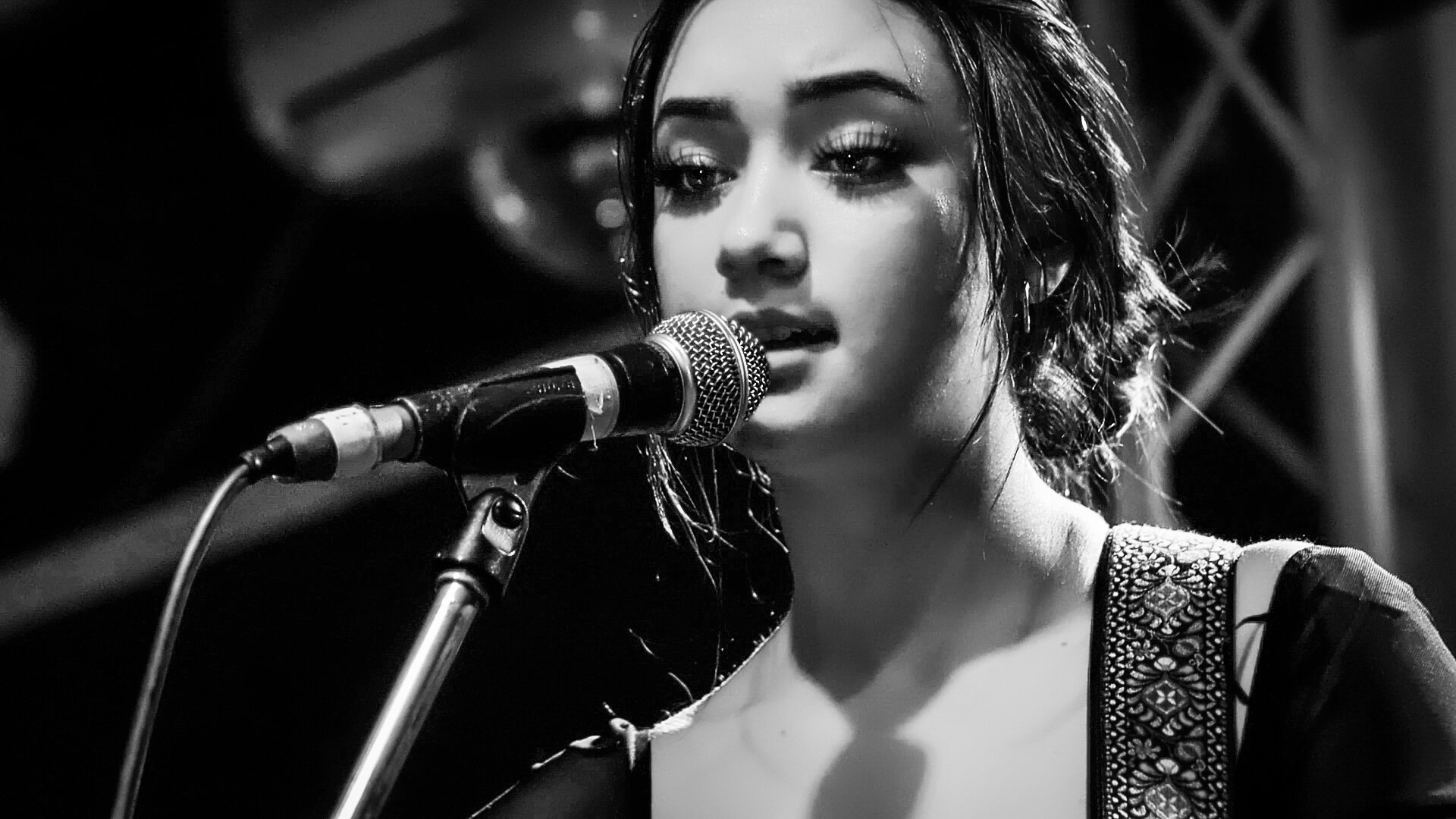 Black and white close-up of a young woman singing into a microphone, eyes closed, with wet hair and a patterned strap on her shoulder.