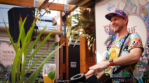 A smiling man wearing a colorful floral shirt and matching hat stands behind a DJ mixer at an outdoor event, with greenery and a mural in the background.