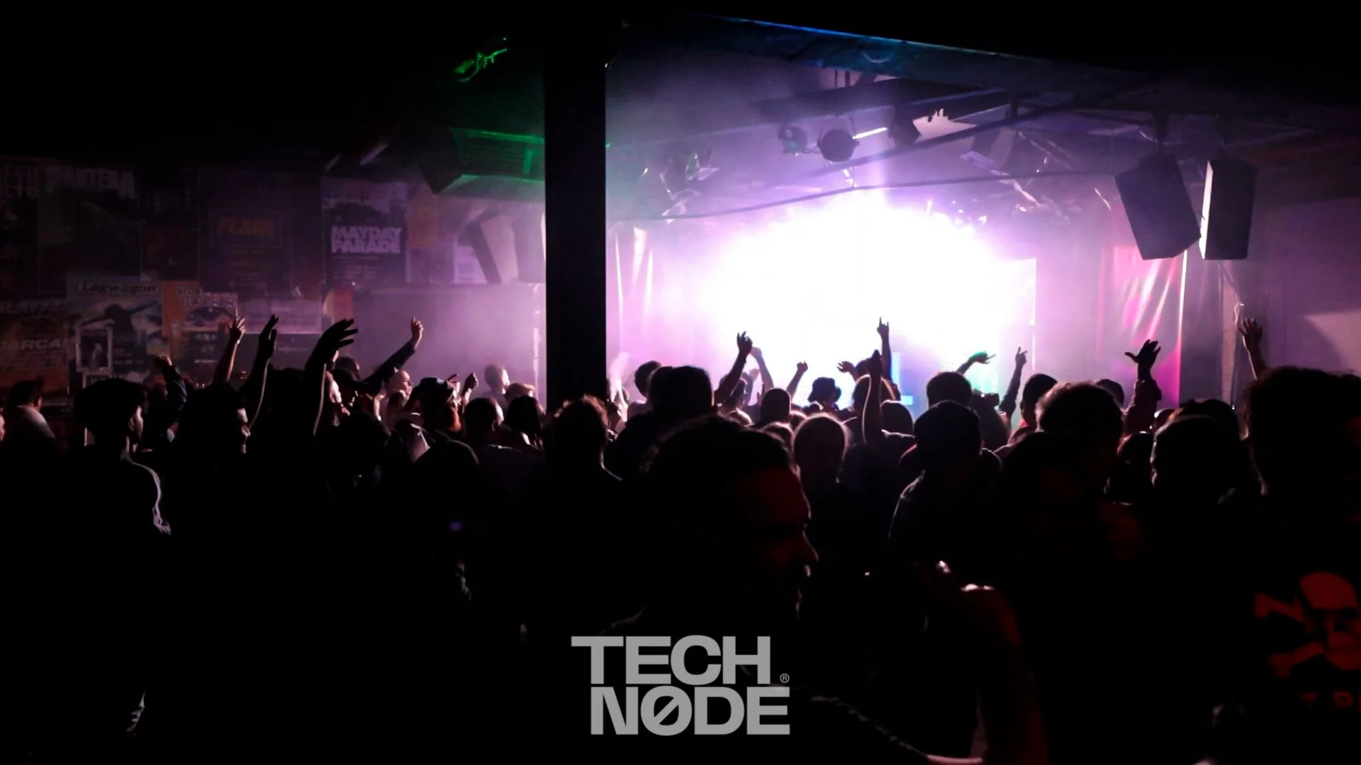 A crowded concert or nightclub scene with silhouettes of people dancing and raising their hands, illuminated by colorful stage lights and a bright white screen in the background.