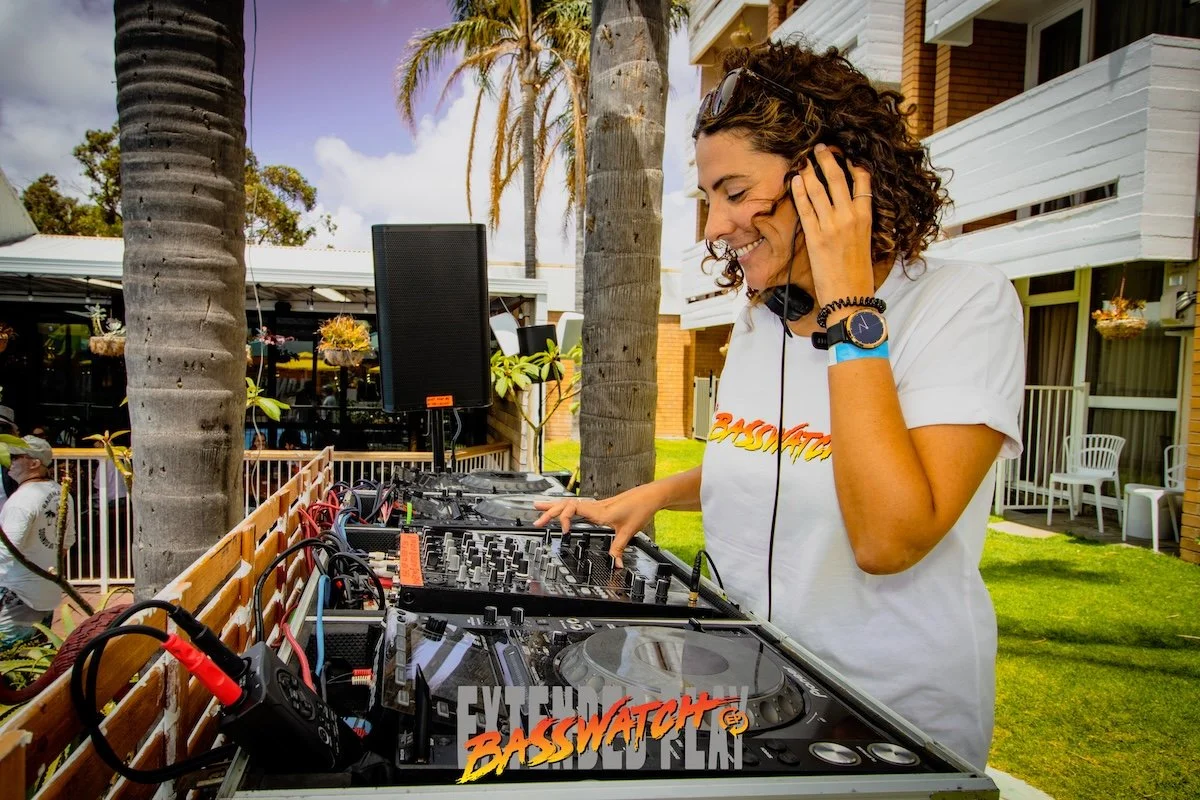 A woman with curly hair, sunglasses on her head, and a white T-shirt smiling while DJing outdoors among palm trees. She is touching a DJ controller with one hand and wearing headphones. The setting is a sunny outdoor area with a building and chairs i
