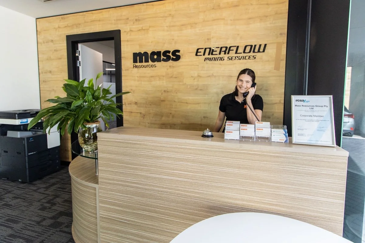 Reception desk with a woman smiling and talking on the phone, three signs on the desk, a framed certificate, a potted plant, a printer, and company logos on a wooden accent wall reading 'mass Resources' and 'EERFLOW MINING SERVICES.'
