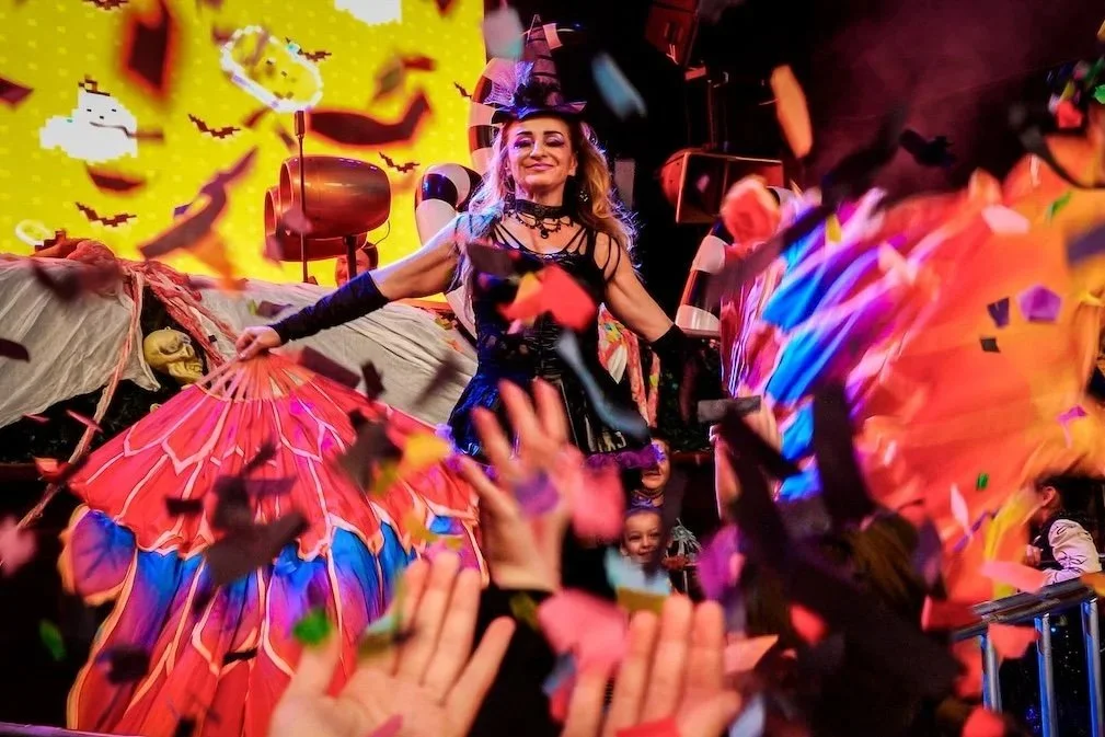 A woman dressed in a witch costume standing on a float at a Halloween parade, with children and colorful decorations around her, and confetti falling from above.