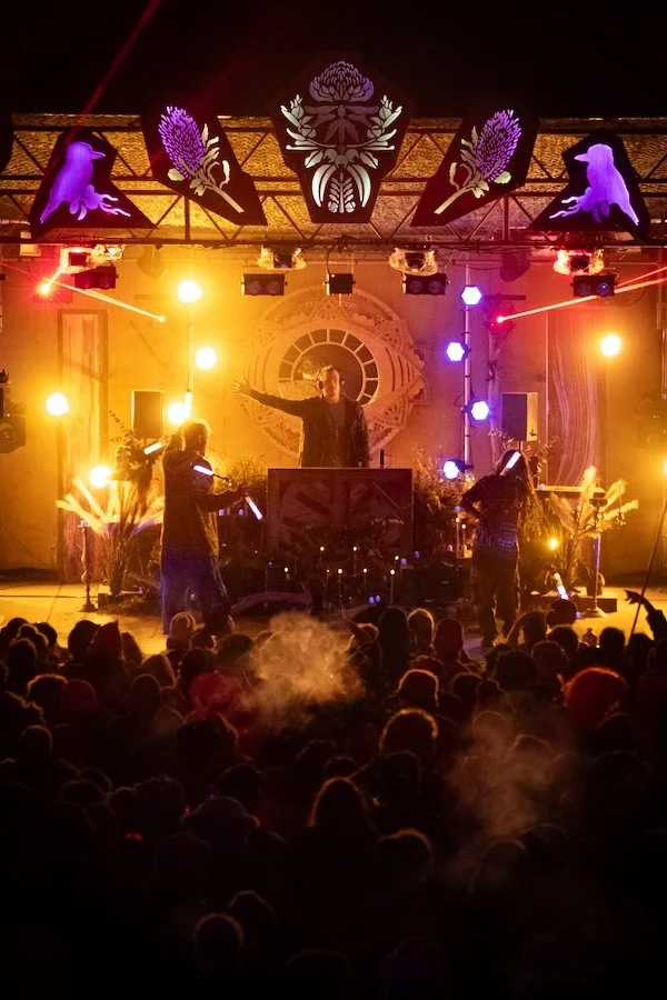 A DJ performs on stage in front of an audience at a concert or event, with colorful stage lighting and decorative elements overhead.