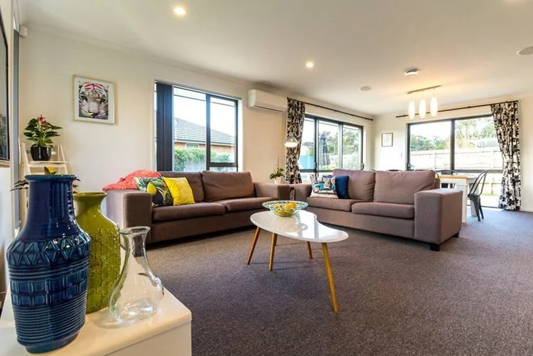 Bright living room with two sofas, a white coffee table, and large windows with curtains. Decorative vases on a nearby table and framed artwork on the wall.