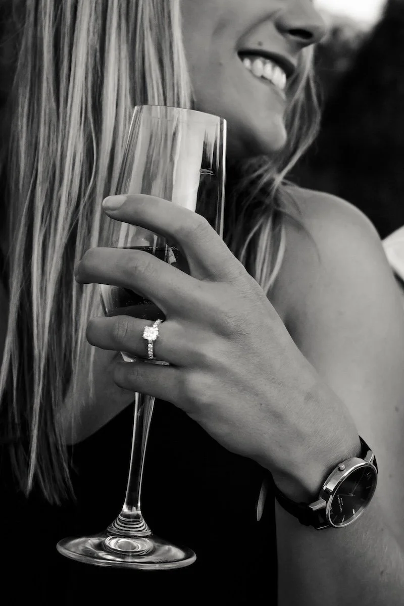A woman smiling and holding a wine glass in her hand, wearing an engagement ring and a wristwatch.