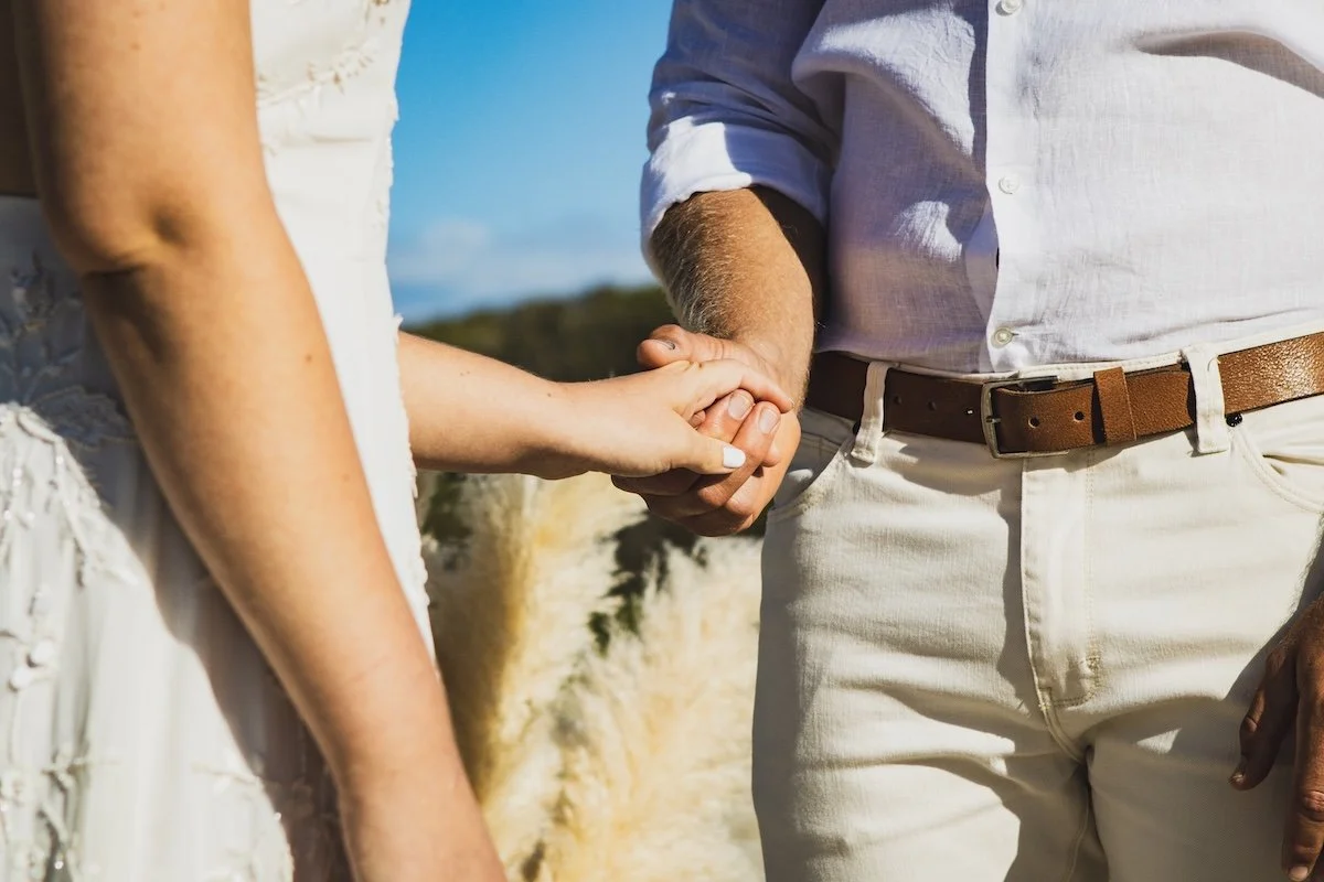 Close-up of a couple holding hands outdoors, with the woman wearing a white dress and the man dressed in a white shirt and beige pants, against a background of cliffs and blue sky.