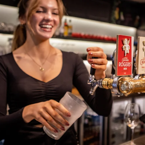 A smiling woman pours beer from a tap into a glass at a bar.