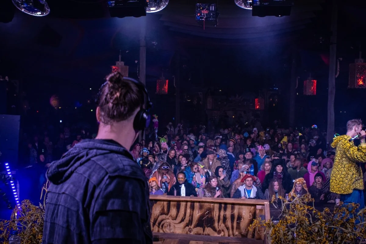 A performer with headphones in a dark venue facing a large audience, which includes people wearing colorful costumes and accessories.