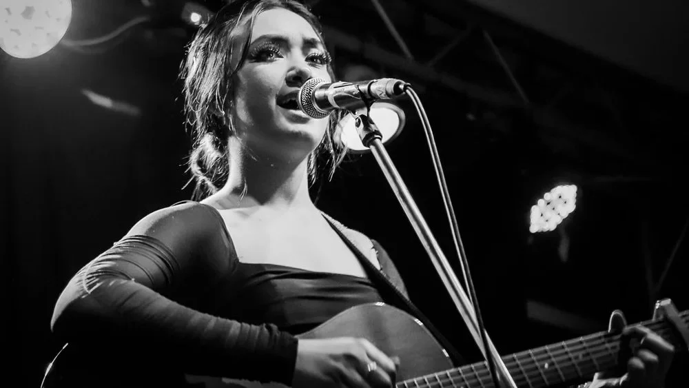 A woman performing on stage with a guitar and singing into a microphone, black and white photo, stage lighting in background.
