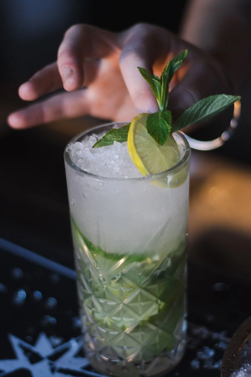 A tall glass cocktail garnished with lemon slices, mint leaves, and ice, being prepared or served.
