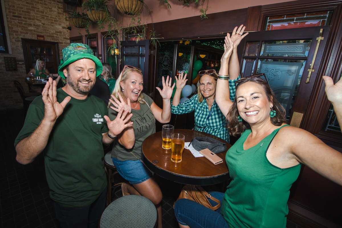 Four people smiling and having fun at a bar or restaurant, raising their hands in celebration, with drinks on the table and a festive atmosphere.