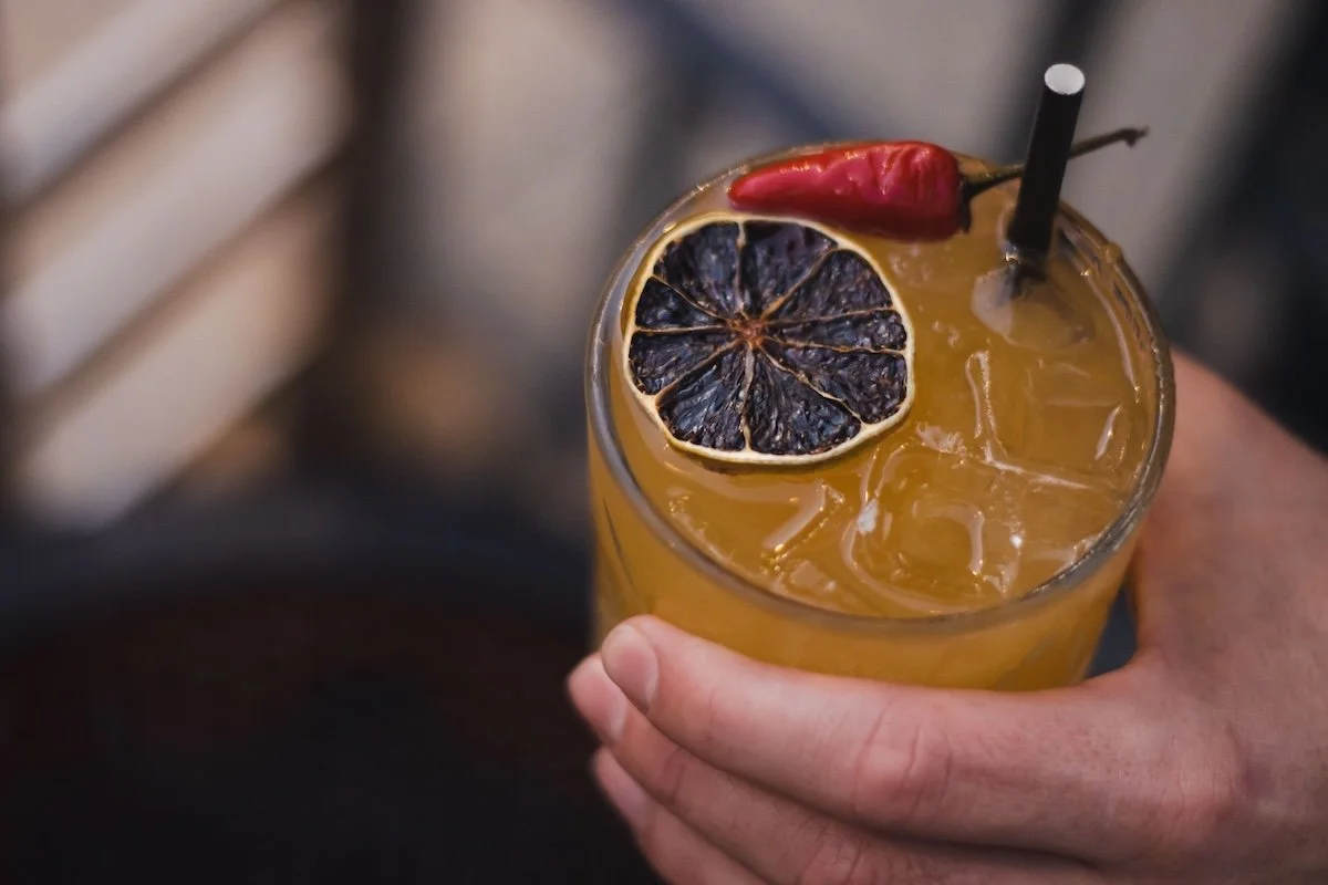 A hand holding a glass of yellow-orange cocktail with ice, garnished with a slice of dried lemon and a red chili pepper, and a black straw.