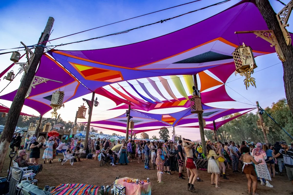 People gathered at a festival under colorful, geometric fabric canopies and hanging lanterns on a dirt field during daytime.