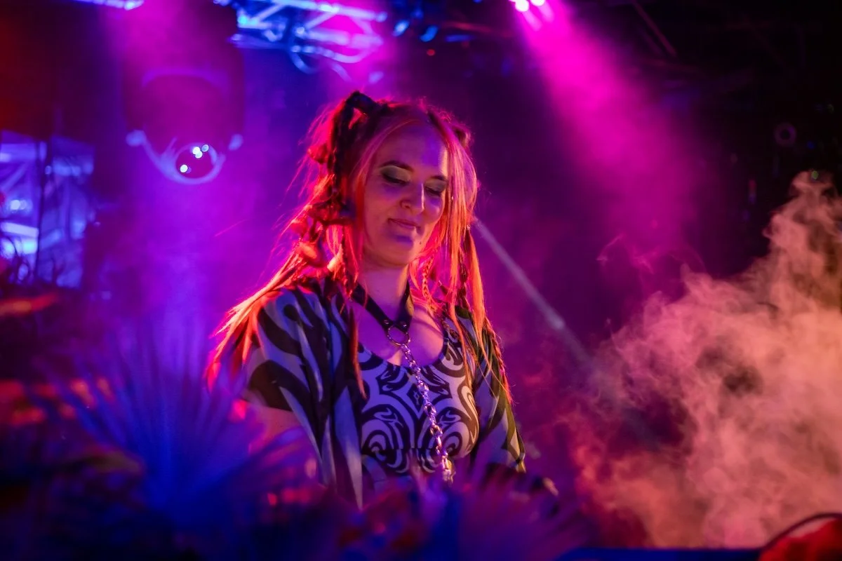 A woman with pink and orange dreadlocks performing as a DJ on stage with purple and pink lighting and smoke effects.