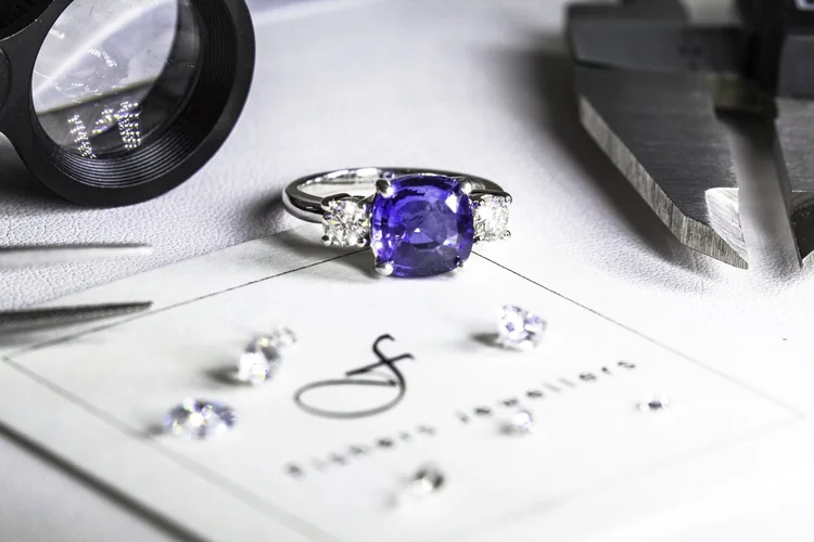 A silver ring with a large blue gemstone and smaller diamonds on either side, surrounded by small loose diamonds, a magnifying glass, and jewelry-making tools on a surface.