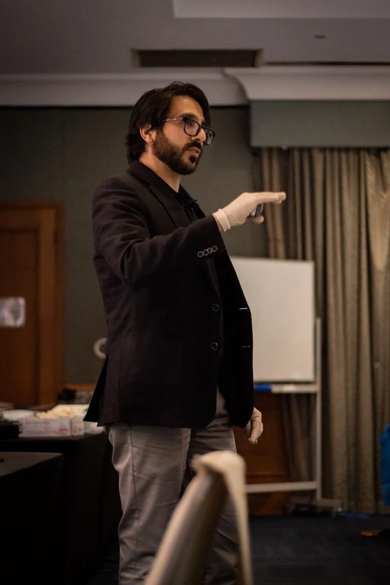A man with dark hair, glasses, and a beard wearing a black blazer and gray pants, standing and gesturing with his right hand in a room with curtains and a whiteboard in the background.