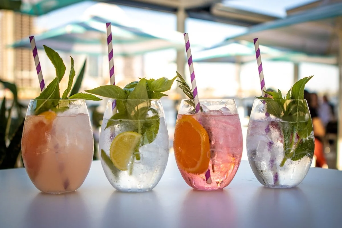 Four glasses with colorful beverages, garnished with mint leaves and lemon slices, on a outdoor patio table with umbrellas in the background.