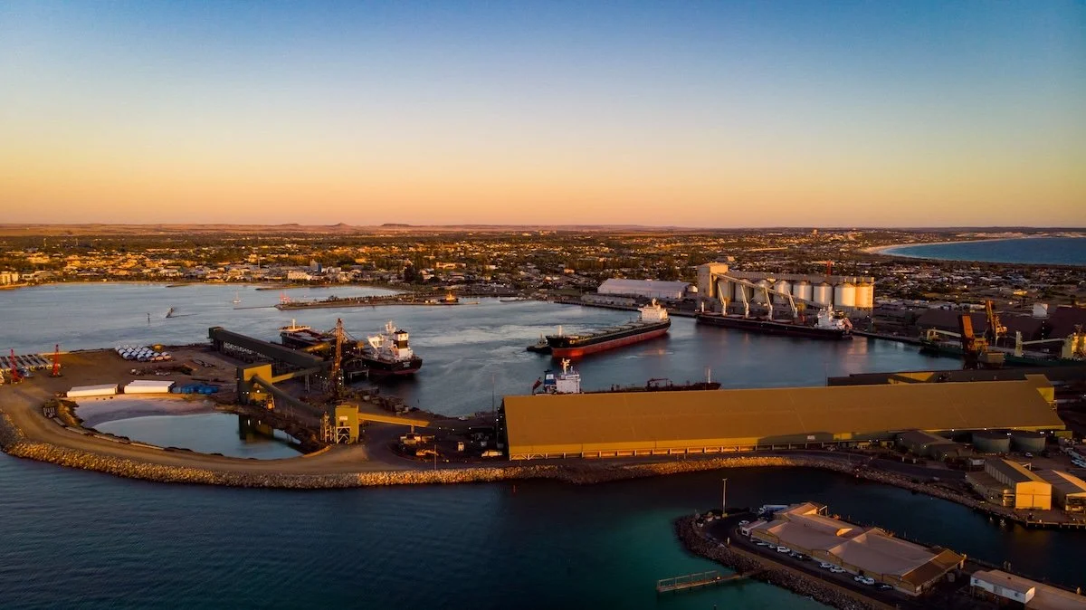 Aerial view of a busy harbor with ships, docks, and industrial warehouses during sunset, near a shoreline and cityscape.