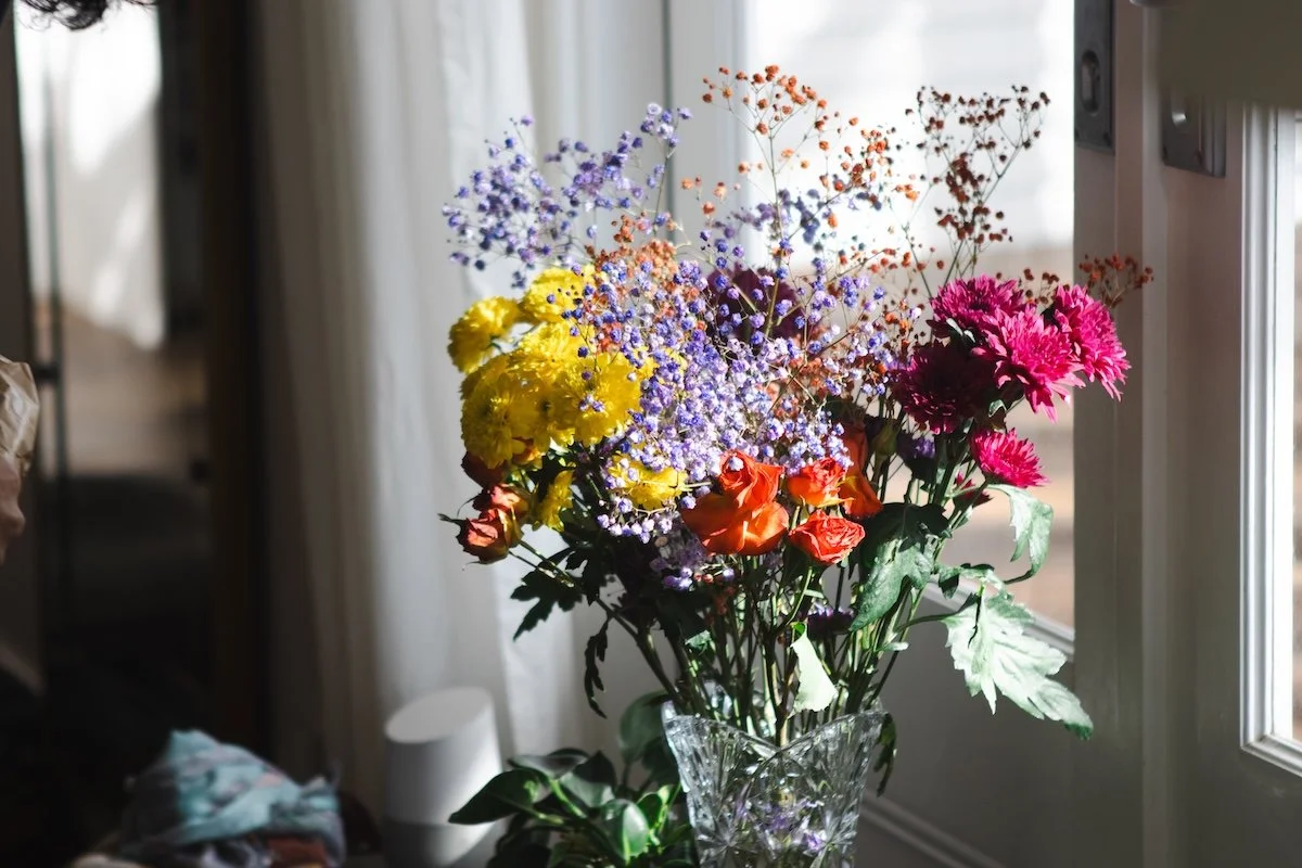 A bouquet of colorful flowers in a clear glass vase placed near a window with sunlight streaming in.