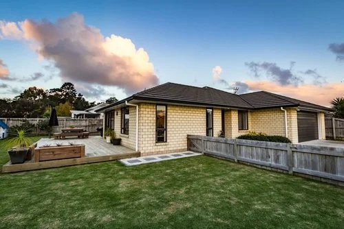 A backyard with a single-story house, a grassy lawn, and a wooden fence, under a partly cloudy sky at sunset.