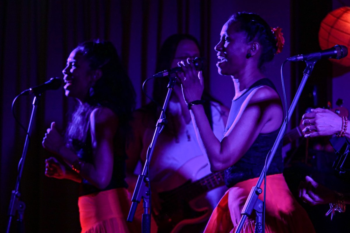 Three women singing on stage with microphones, colorful lighting, and a red paper lantern in the background.