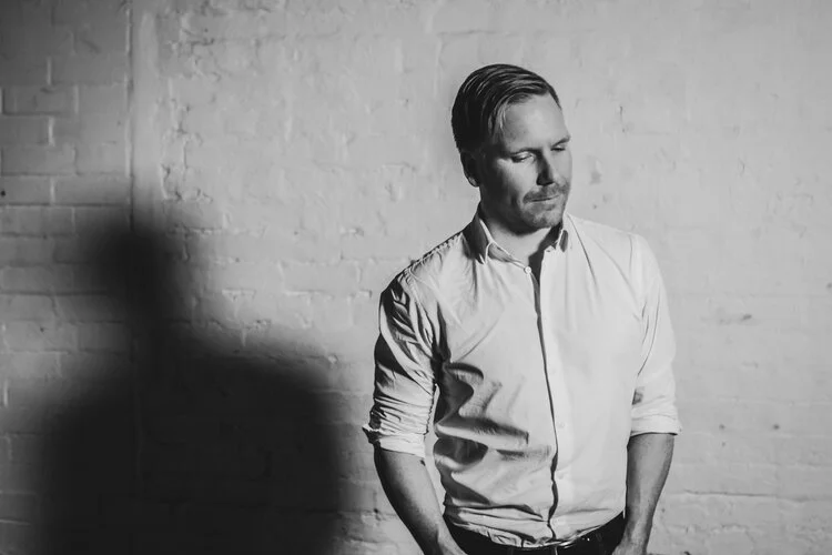 A man with slicked-back hair and a beard, wearing a light-colored, long-sleeve, button-up shirt with rolled-up sleeves, stands against a brick wall. He has his hands in his pockets and appears to be looking down thoughtfully.