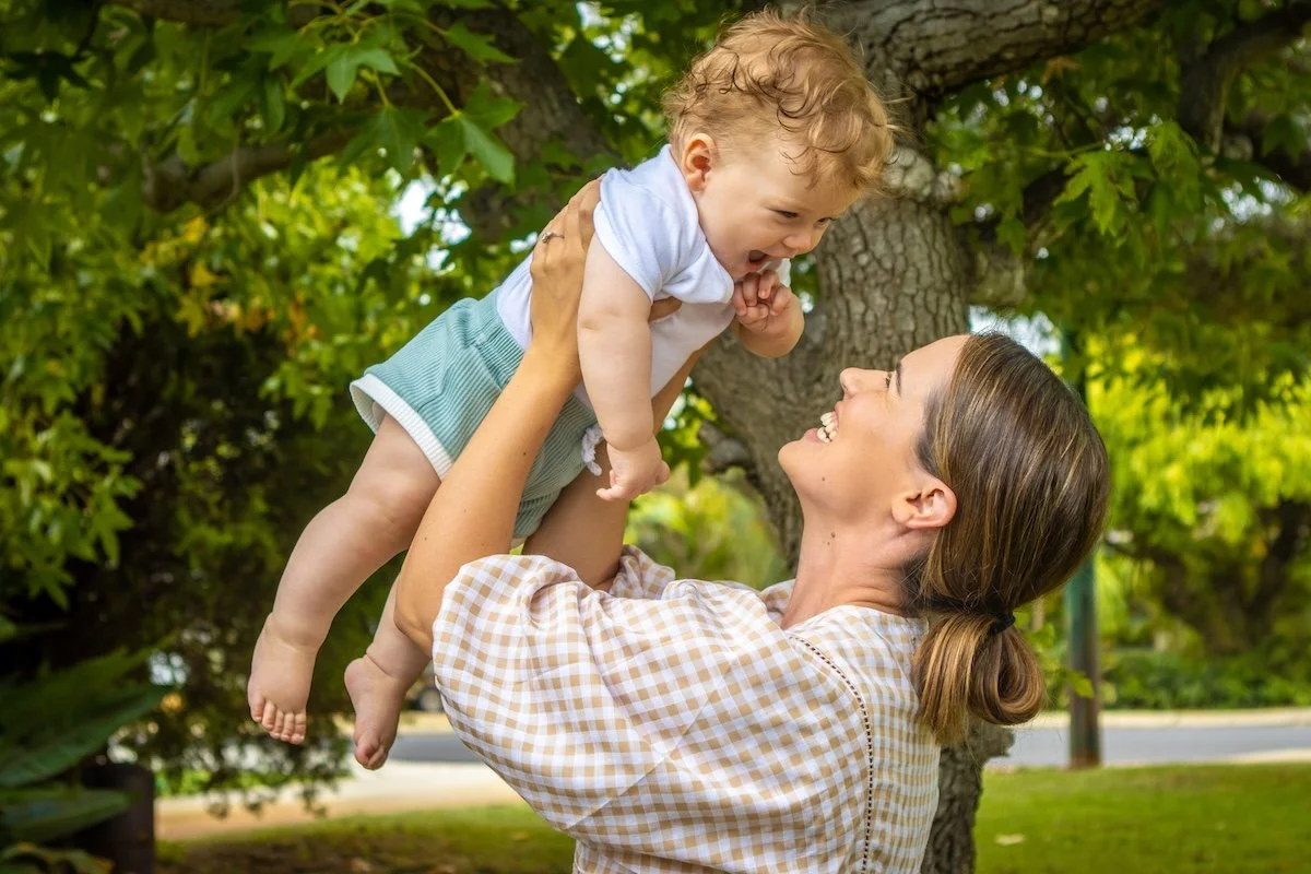 A woman lifting a smiling young child in the air outdoors under a tree.