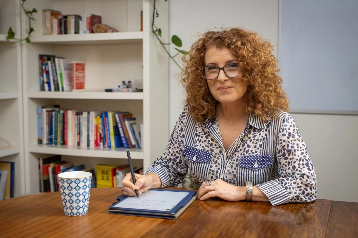 A woman with curly red hair and glasses sitting at a wooden table, writing on a digital tablet with a stylus. There is a blue and white patterned mug on the table, and a white bookshelf with books and decorations in the background.