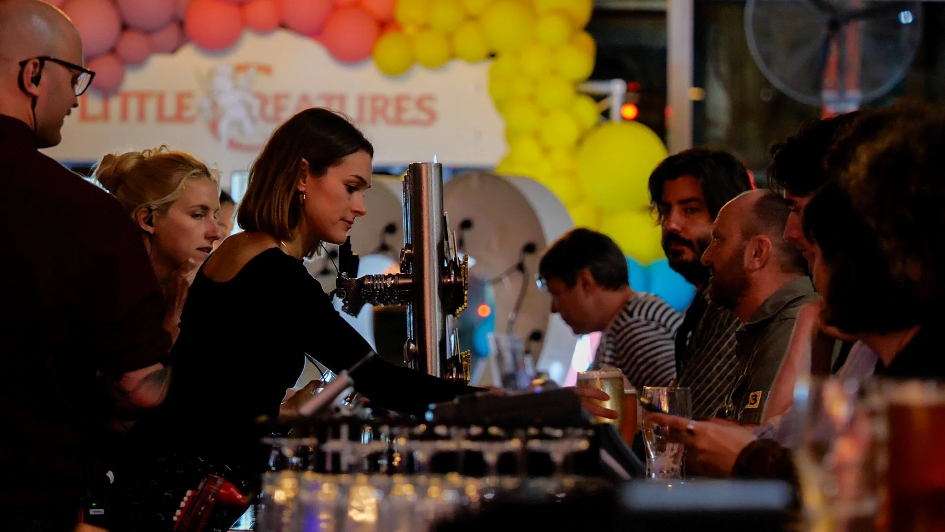 A woman serves drinks at a bar counter during a lively social event, with a group of people seated on the other side, and colorful balloons and a sign that reads 'LITTLE TREASURES' in the background.