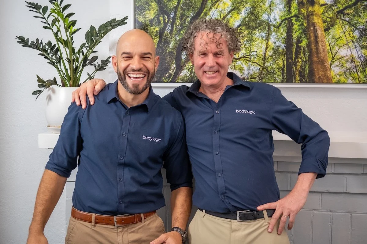 Two men wearing matching blue shirts with the logo 'bodylogic' on them, smiling and posing together with one man’s arm around the other.