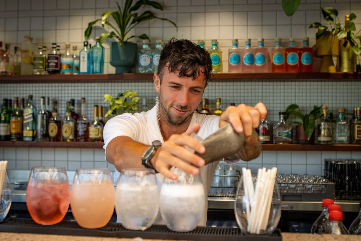 A bartender preparing drinks behind a bar with various bottles and plants on shelves.
