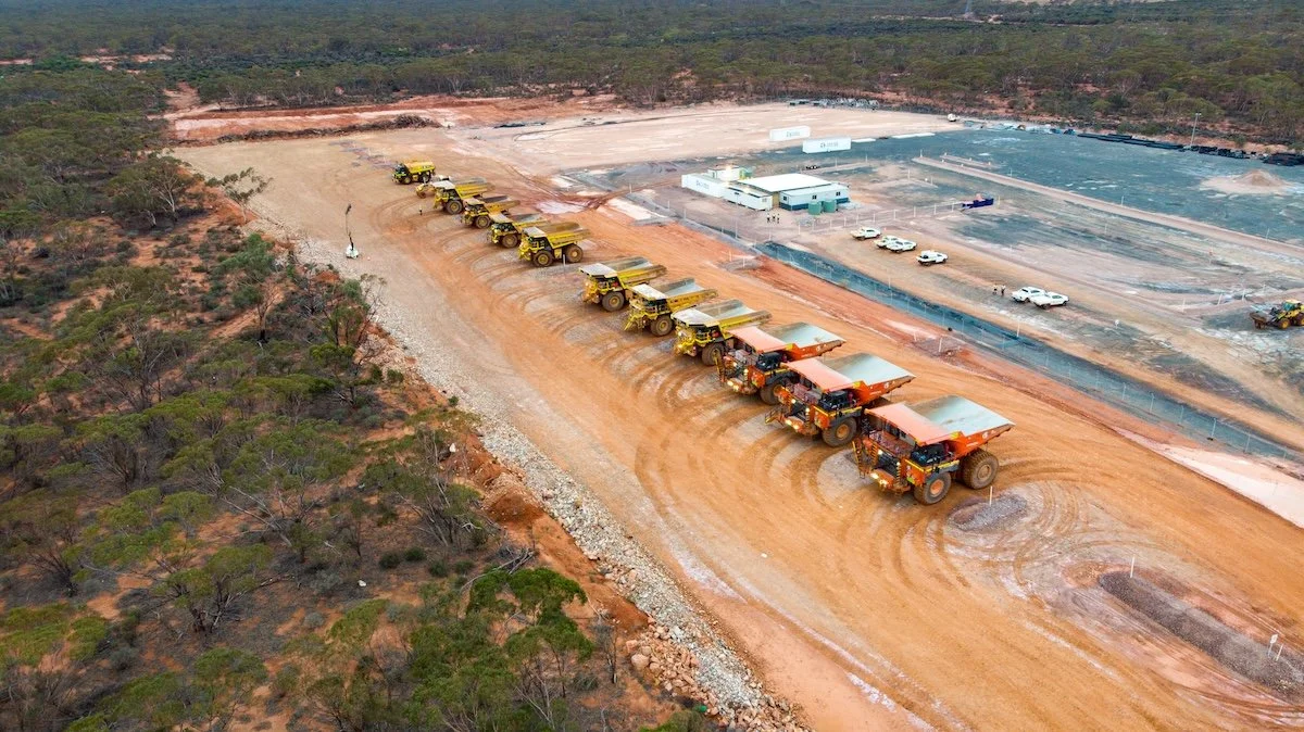 Aerial view of a construction site with a row of large dump trucks on a dirt road, surrounded by trees and some construction equipment, with a fenced area and parked cars in the background.