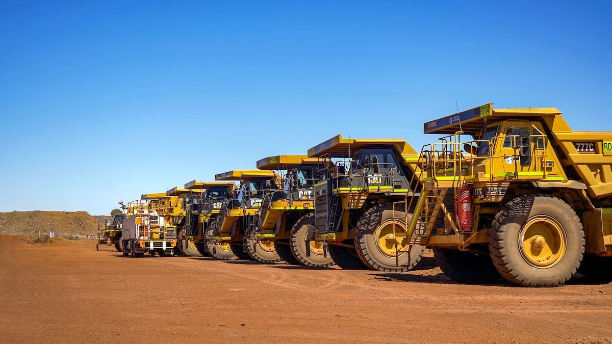 A row of yellow Caterpillar heavy-duty dump trucks parked on reddish dirt under a clear blue sky.