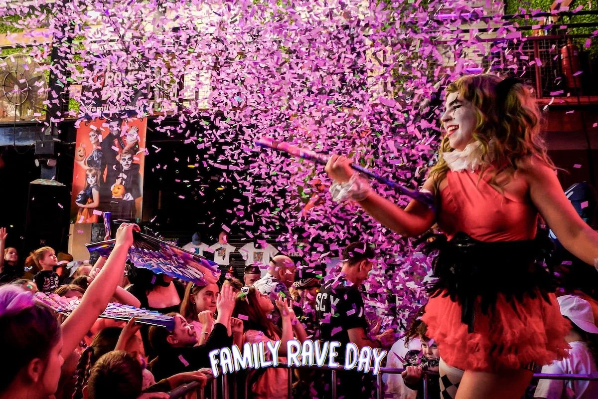 A lively family rave party with confetti falling, featuring a performer dressed as a clown or carnival character with face paint, wearing a red and black costume, entertaining children and adults.