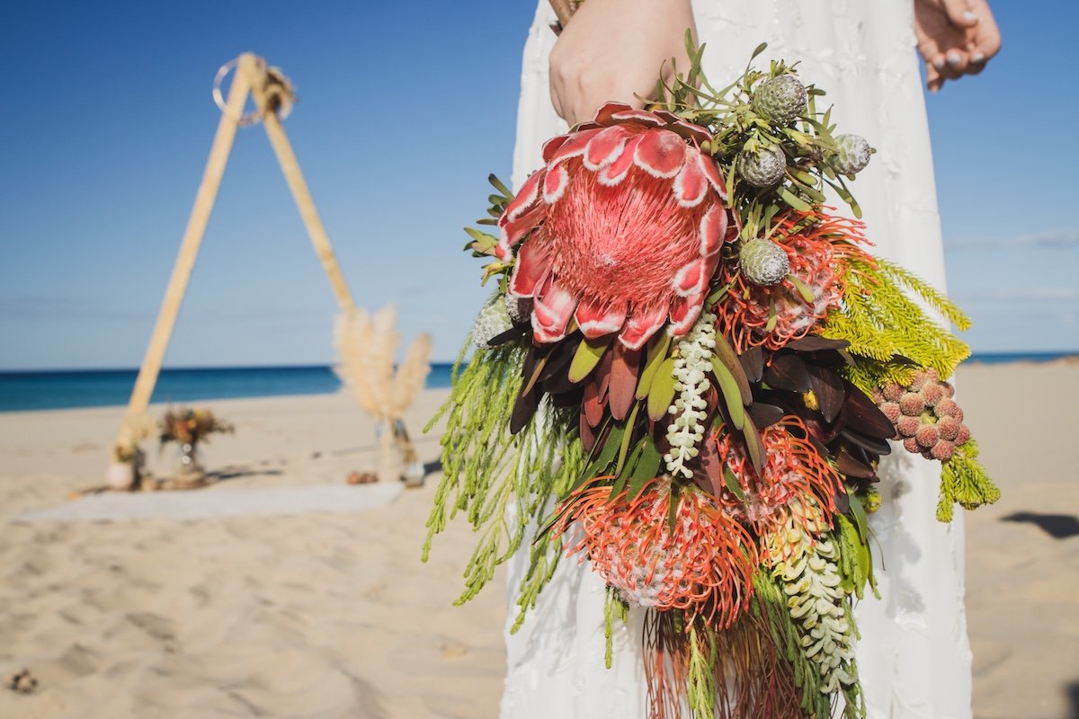 Person holding a tropical flower bouquet on a beach with an arch in the background for a wedding or special event.