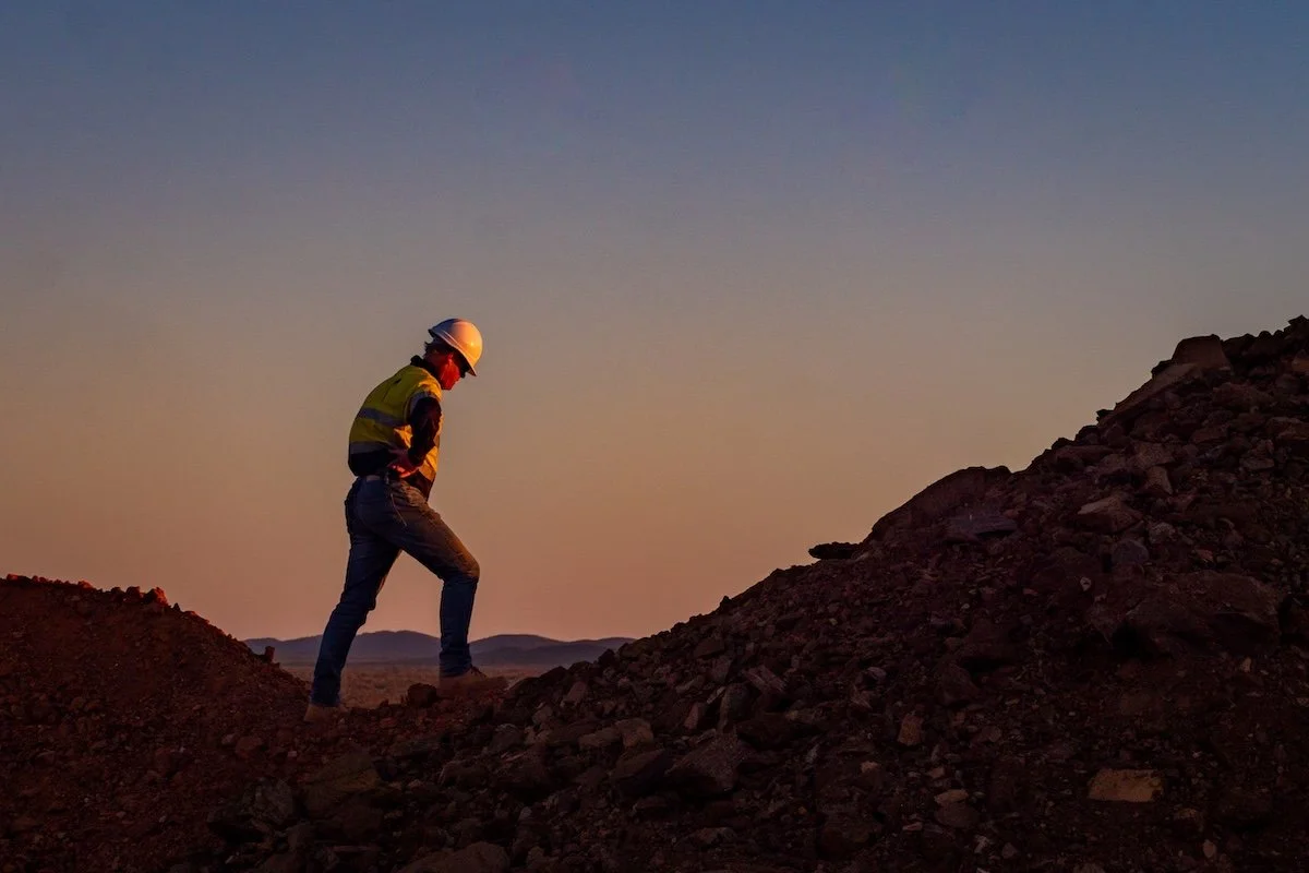 A construction worker wearing a yellow safety vest and white hard hat inspecting a pile of dirt or rocks at sunset.