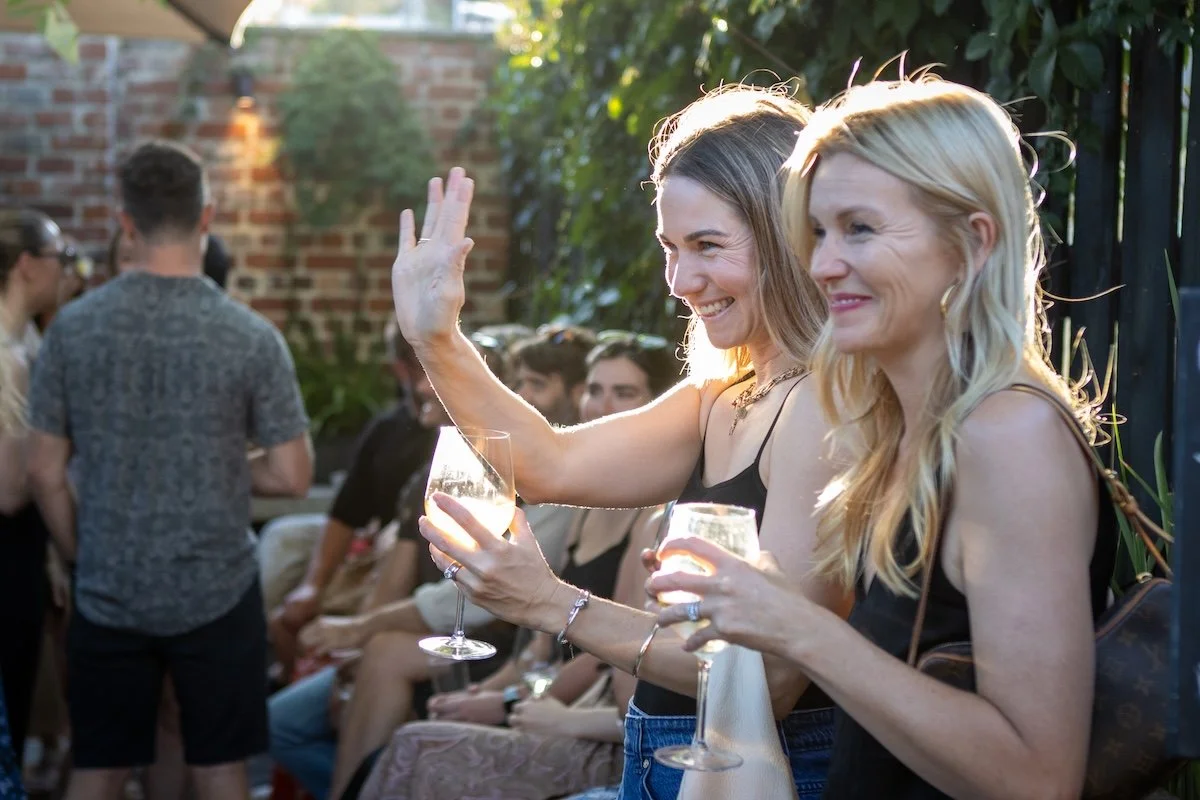 Two women smiling, one waving, holding wine glasses at an outdoor party with other guests and greenery in the background.