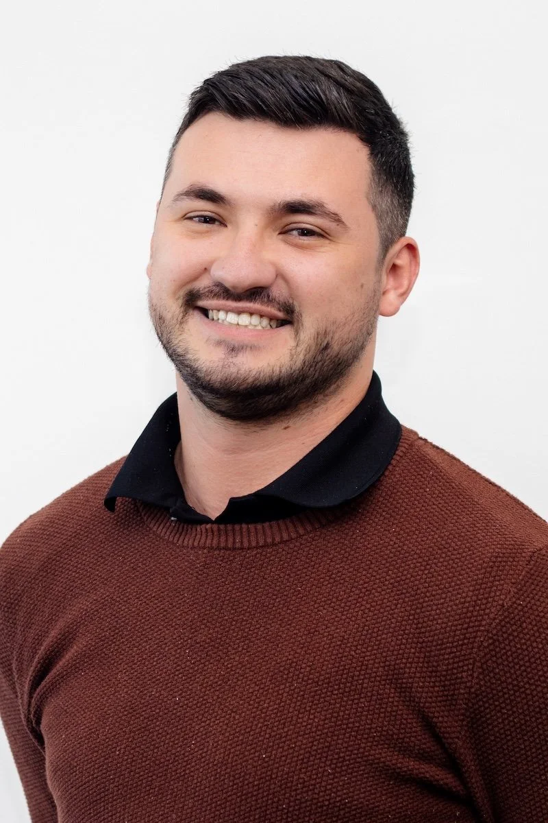 A man smiling with short dark hair and a beard, wearing a black collared shirt and a brown sweater, standing against a white background.